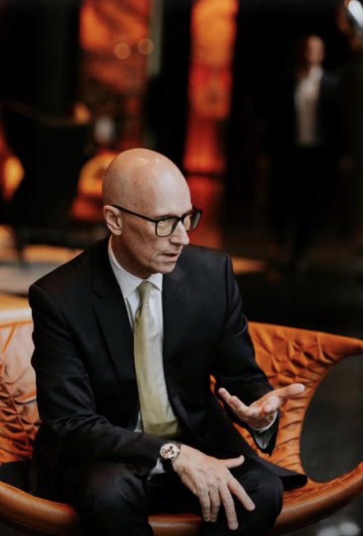 Perry Offer, seated in conversation, wearing a dark suit and glasses.